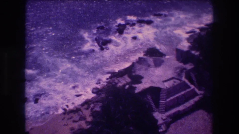 8mm home movie frame from 1982 Acapulco showing turbulent surf breaking over rocks from an elevated vantage, with dark vegetation and a stepped building visible at right.