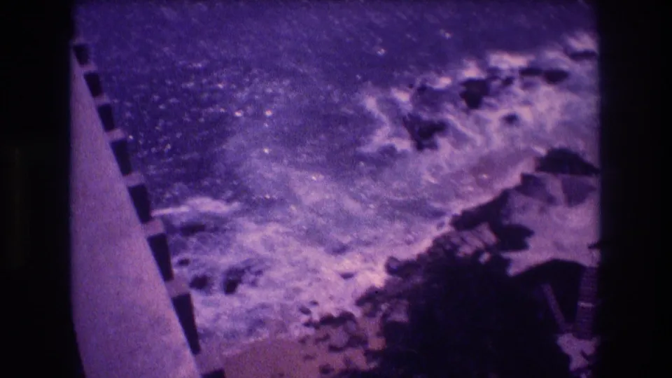 8mm home movie frame from 1982 Acapulco showing waves breaking on rocks directly below a bridge overlook, concrete parapet edge visible at lower left.