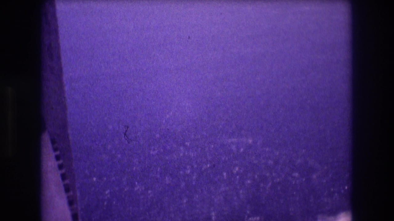 Opening frame of a 1982 Acapulco home movie with deep purple dye shift, film-perforation strip on the left edge, and a grain-heavy ocean expanse filling the lower portion.