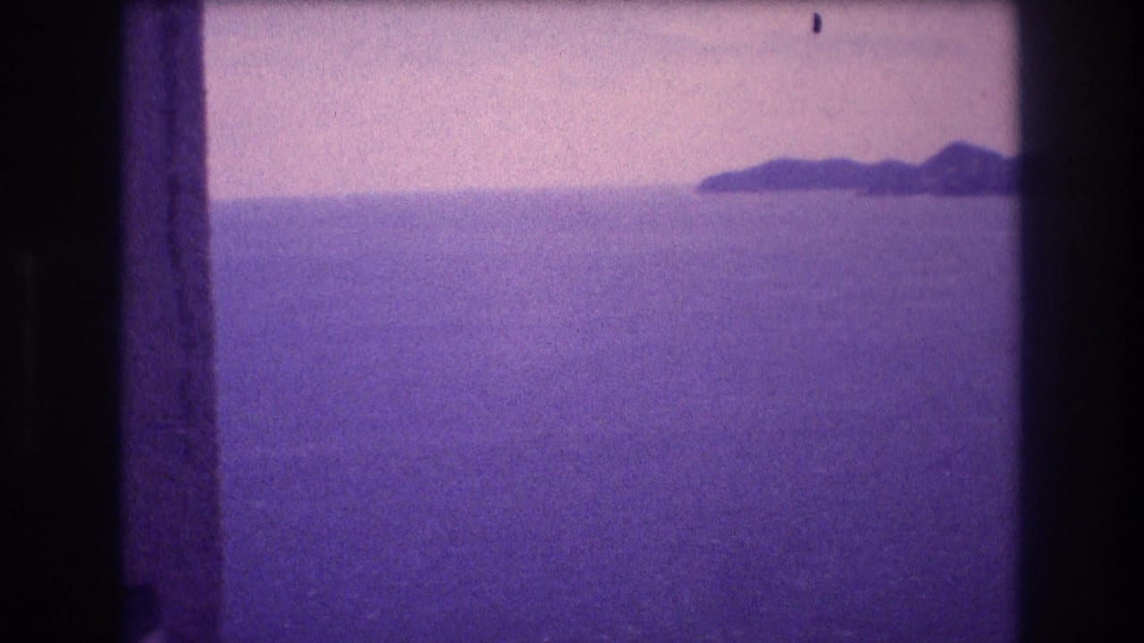 Mid-pan frame from a 1982 Acapulco home movie; an island silhouette emerges in the upper-right horizon over a wide purple-cast ocean beneath a pale, hazy sky.