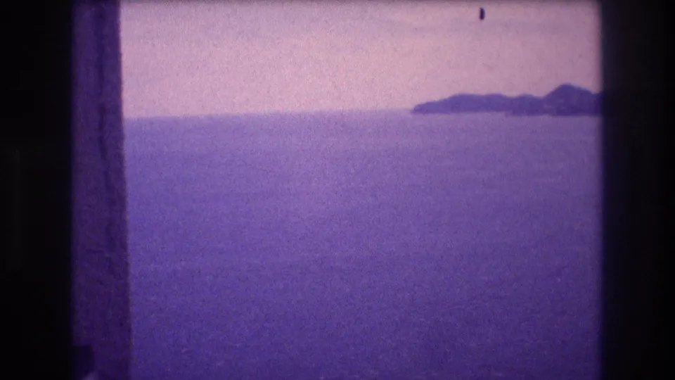 Mid-pan frame from a 1982 Acapulco home movie; an island silhouette emerges in the upper-right horizon over a wide purple-cast ocean beneath a pale, hazy sky.