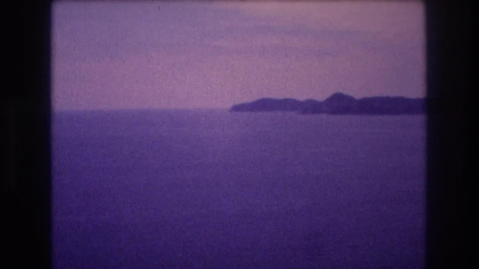 Stabilized view from a 1982 Acapulco home movie showing a low multi-peaked island silhouette centered on the ocean horizon under an overcast sky with strong purple dye shift.