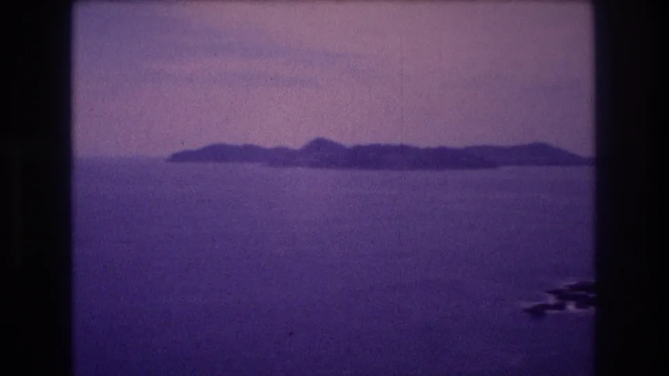 Closing frame of a 1982 Acapulco home movie; an island ridgeline sits on the ocean horizon beneath a pale hazy sky with strong magenta-purple film dye shift and visible grain.