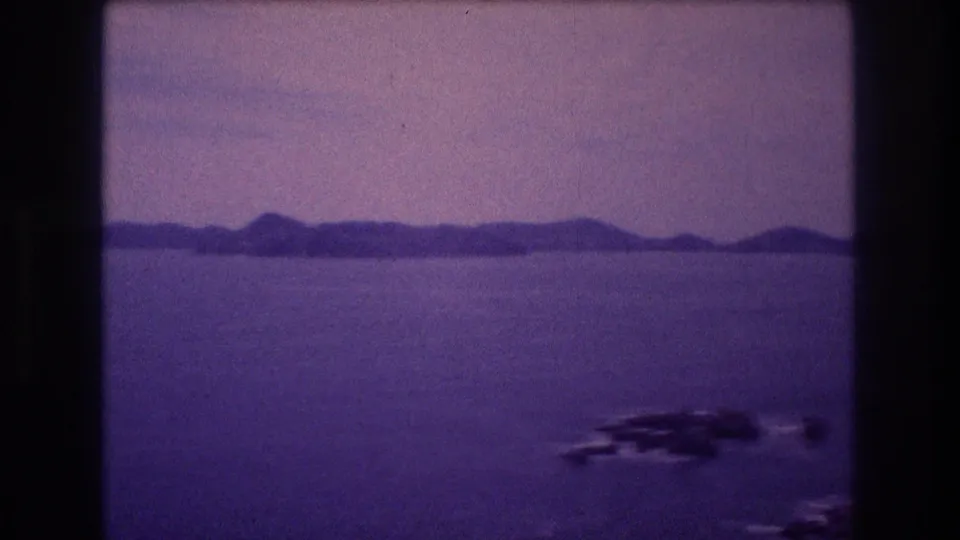 Elevated view over a bay identified as Acapulco circa 1982, showing calm water, distant headlands, and rocky outcroppings on 8mm home movie film.