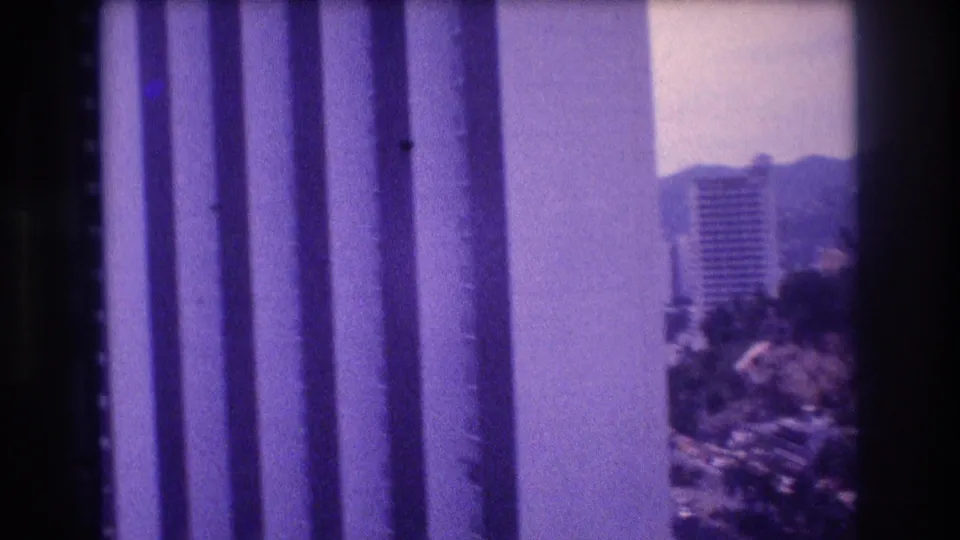 Shot through a balcony or window railing in what appears to be Acapulco circa 1982, revealing a tall high-rise building and hillside vegetation on 8mm film.