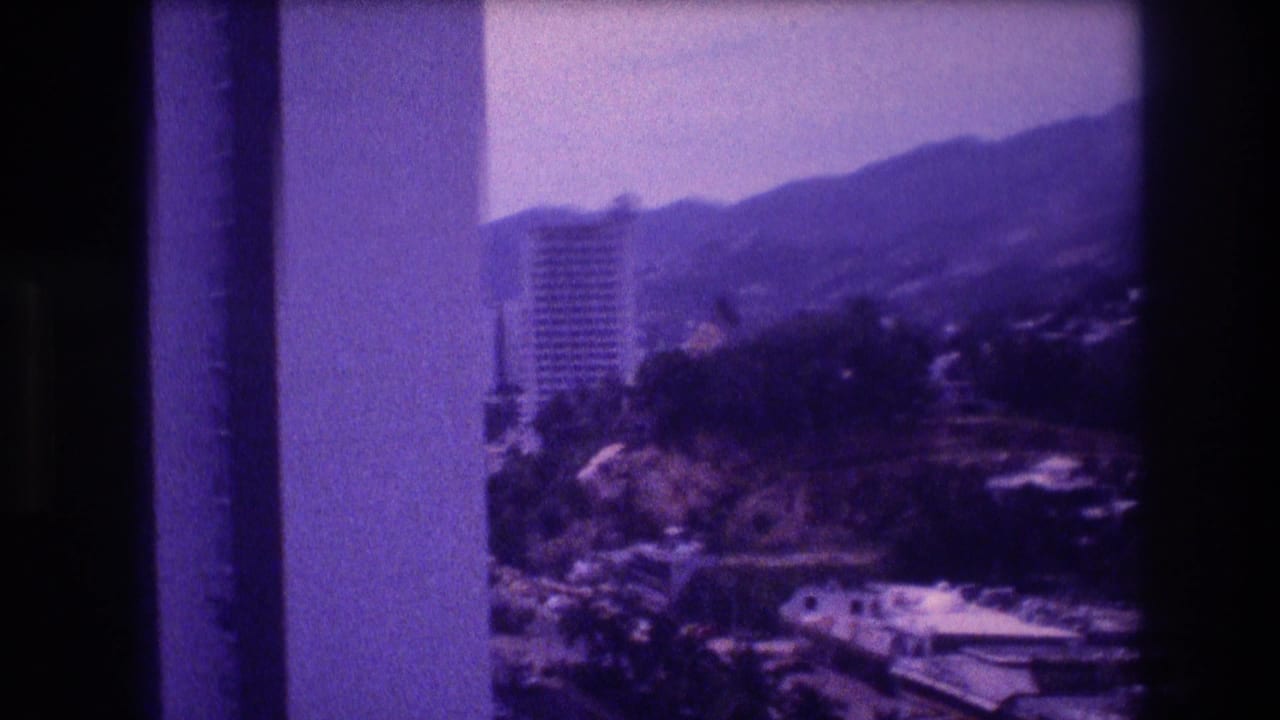Elevated 8mm home-movie frame from Acapulco, Mexico, c.1982 — tall high-rise among treetops, hillside buildings, and mountain ridgeline in the background.