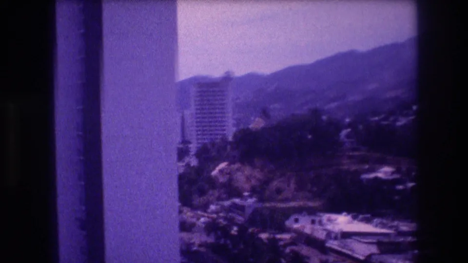 Elevated 8mm home-movie frame from Acapulco, Mexico, c.1982 — tall high-rise among treetops, hillside buildings, and mountain ridgeline in the background.