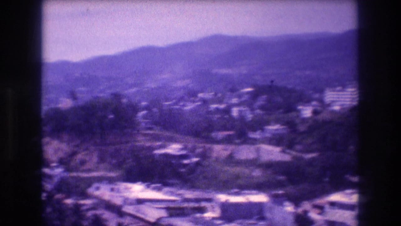 8mm home-movie frame from Acapulco, Mexico, c.1982 — white residential buildings on forested hillsides beneath a hazy mountain ridgeline.