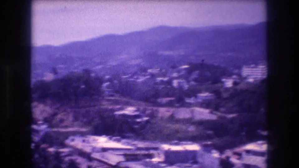 8mm home-movie frame from Acapulco, Mexico, c.1982 — white residential buildings on forested hillsides beneath a hazy mountain ridgeline.