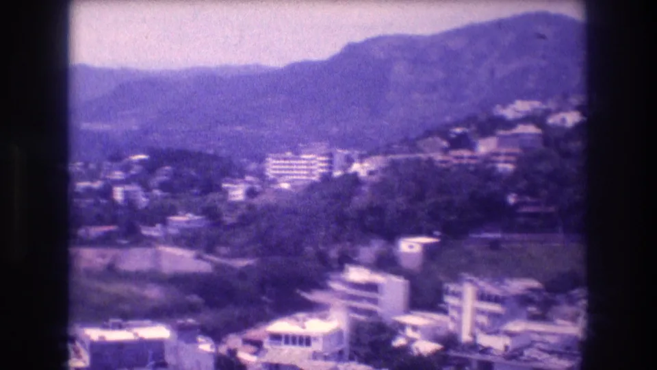 8mm panoramic home-movie frame from Acapulco, Mexico, c.1982 — flat-roofed low-rise buildings in the foreground, hillside residential development, and a mountain range behind.