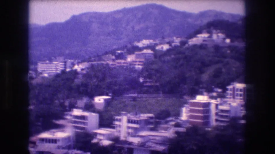 8mm home-movie pan frame from Acapulco, Mexico, c.1982 — low-rise buildings and hillside residential development backed by a forested mountain range.