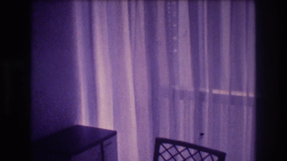 8mm home-movie frame at pan end showing sheer draped curtains, a small dark-surfaced desk, and a lattice-back chair in what appears to be an Acapulco apartment, 1982