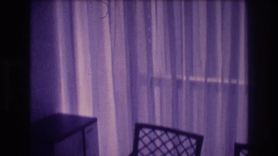 1982 8mm home movie interior: sheer curtains dominate the frame with a lattice-back chair at lower center, shot in Acapulco, Mexico with pronounced purple film color shift.