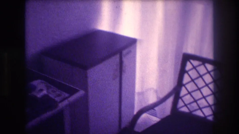 Mid-pan 8mm frame from 1982 Acapulco showing white nightstand, lattice chair, and backlit sheer curtains in a vacation room interior.