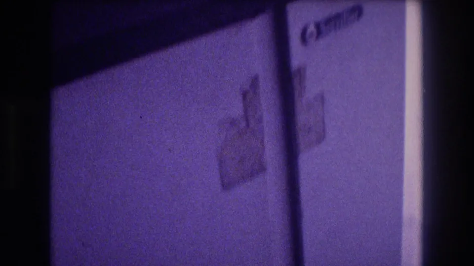 White compact refrigerator exterior showing an adhesive sticker or label, door partially in frame, heavy purple Super 8 color cast, indoor scene circa 1982.