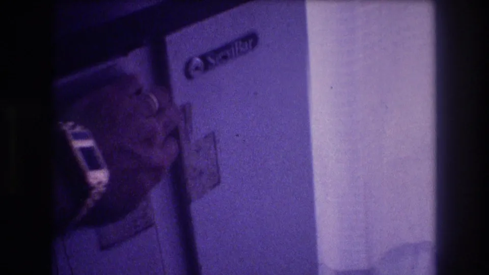 Man's head partially in frame at lower left, open compact refrigerator with a brand label visible, low-light indoor scene, Super 8 home movie circa 1982 Acapulco Mexico.