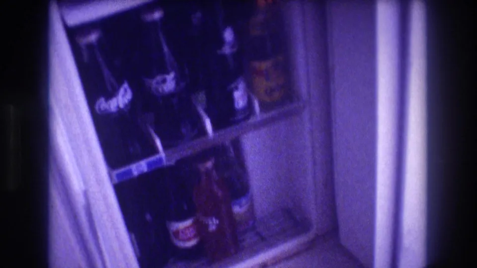 8mm home movie frame from circa 1982 showing the interior of an open refrigerator stocked with Coca-Cola and other bottled beverages, with a heavy purple film cast.