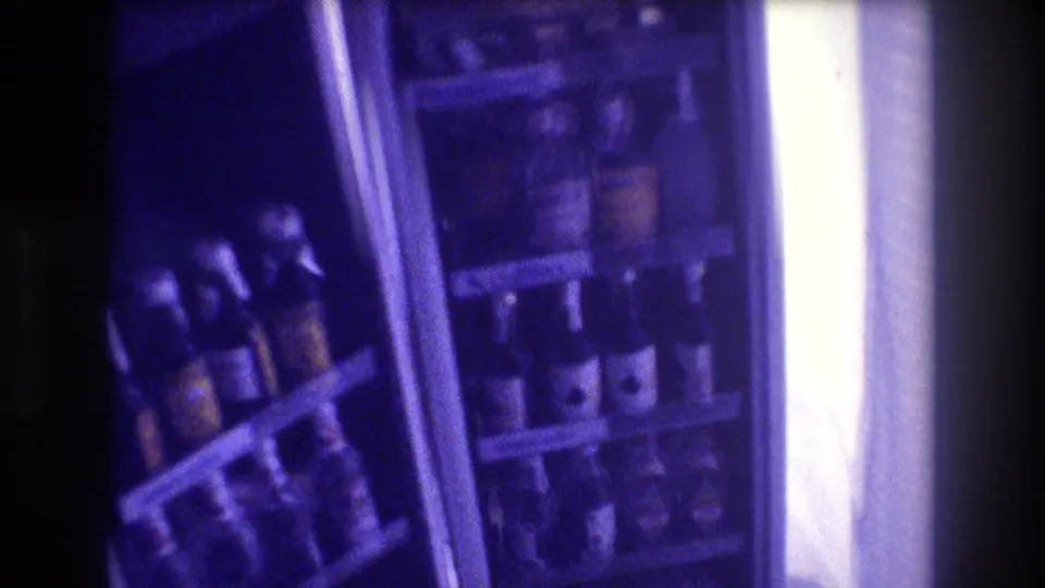 8mm home movie frame circa 1982 showing a second refrigerator fully stocked with multiple shelves of assorted bottled and canned drinks, distinct purple film cast.