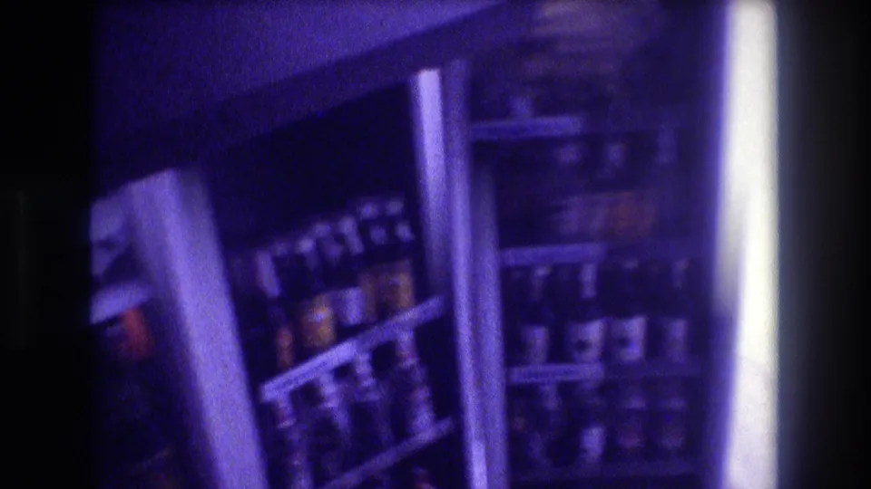 8mm home movie frame showing a densely stocked refrigerator with bottles and cans, interior light and purple color cast, circa 1982.