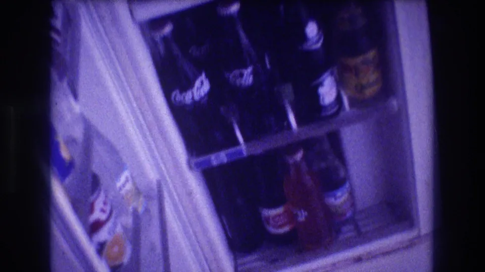 A closer 8mm view of the refrigerator door and shelves shows multiple glass soda bottles, including visible Coca-Cola branding, in a private interior.