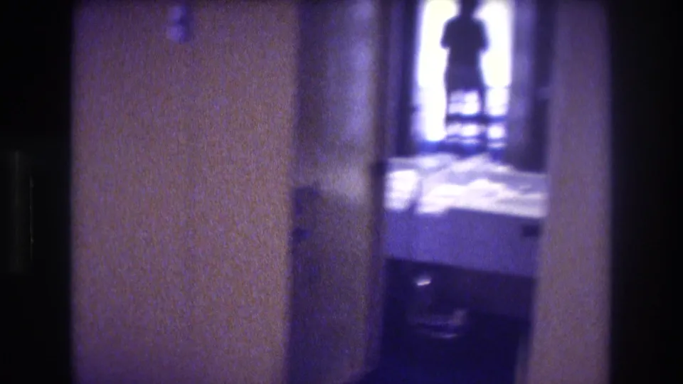 Final 8mm frame of a pan — dim interior hallway with tan walls and a backlit adult silhouette in a bright doorway opening, Acapulco circa 1982.