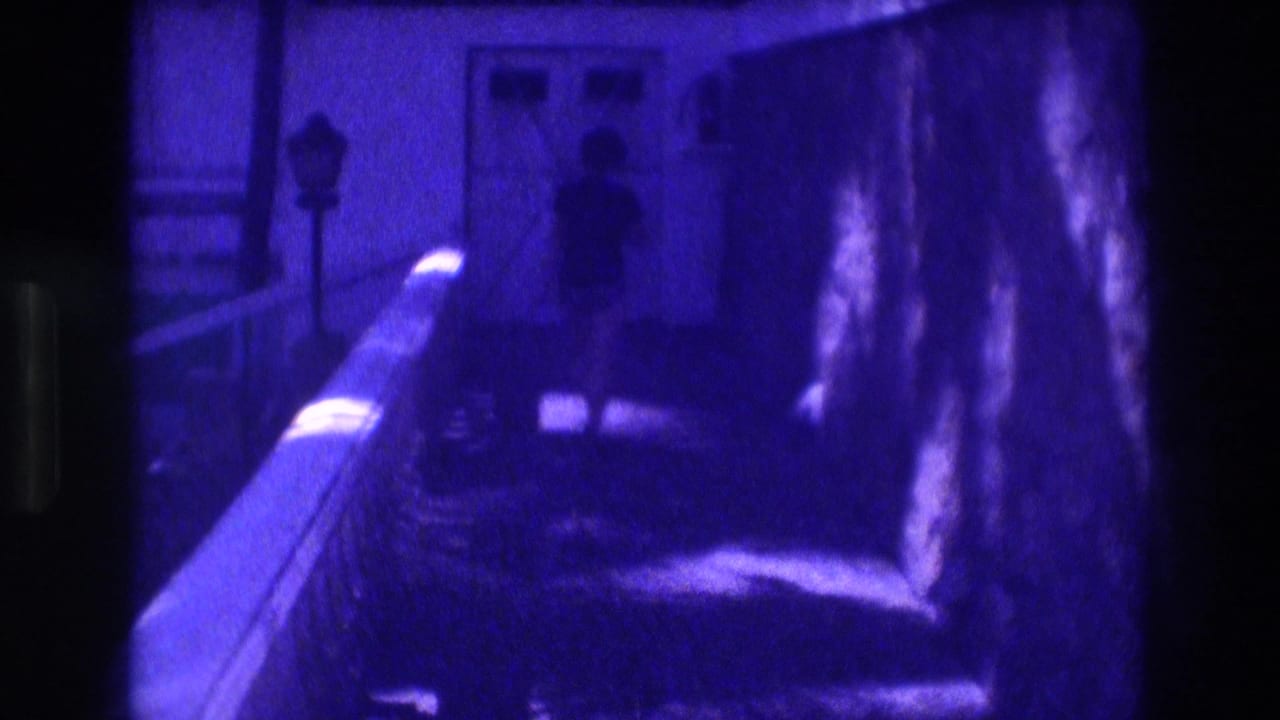 Balcony corridor with diagonal railing and purple-blue tint; silhouetted figure walking; lamp post and tropical vegetation visible.