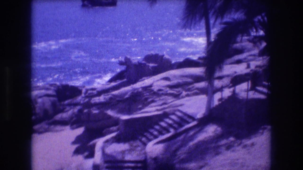 Aged 8mm frame showing a rocky Pacific shoreline at Acapulco, Mexico, circa 1982, with stone steps, a palm tree, and crashing surf under a bright sky.