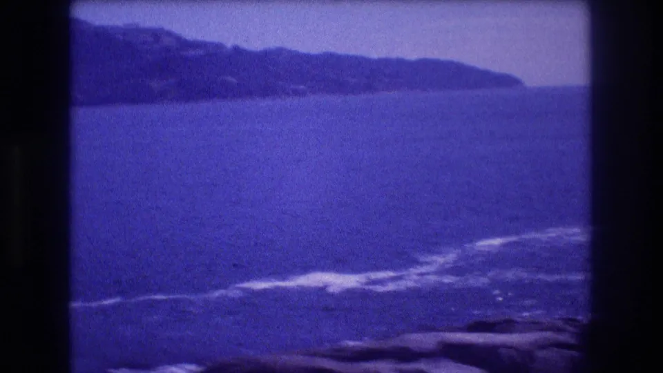 Wide 8mm pan frame showing an open Pacific bay at Acapulco, Mexico, circa 1982, with a distant headland, breaking surf on foreground rocks, and deep purple-blue tones.