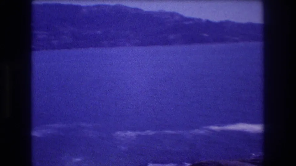Final 8mm frame of an Acapulco bay, circa 1982, showing a prominent headland silhouette across open water, with rocks and surf in the foreground and a deep purple-blue color shift.
