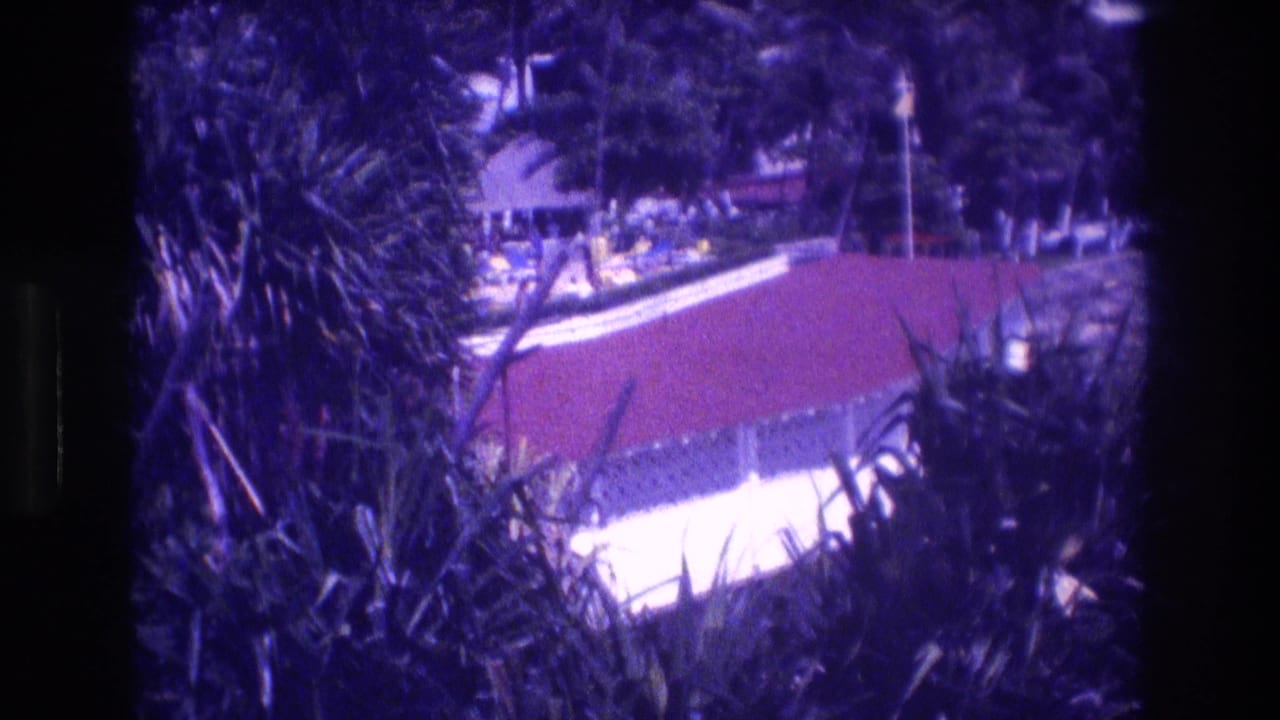 Panning shot through tropical foliage toward the red-roofed resort building identified as Acapulco, Mexico, 1982; white structural element and trees visible beyond the plants.