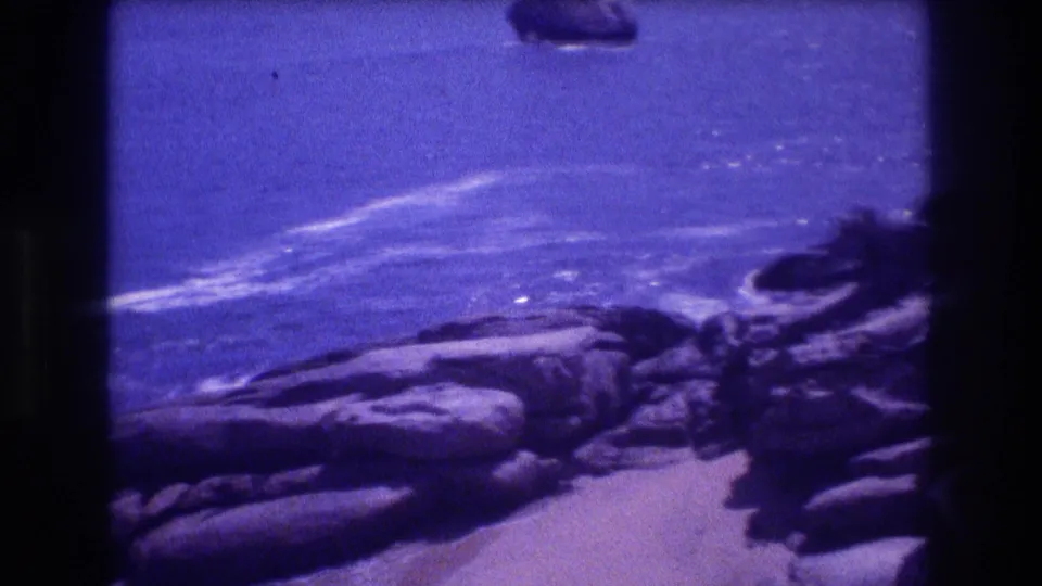 8mm home-movie frame of a rocky beach with white wave crests and blue ocean beyond; purple dye-shift and grain typical of aged 1980s home-movie stock, likely Acapulco, Mexico.
