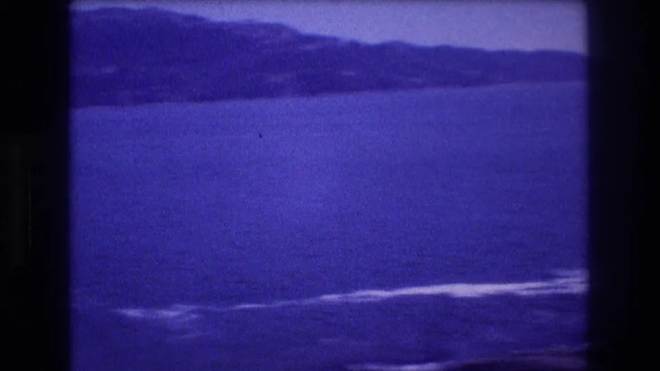 Mid-pan 8mm home-movie frame from Acapulco, Mexico, circa 1982: open bay with surf, rocky elements receding, and hazy foothills under a flat sky.