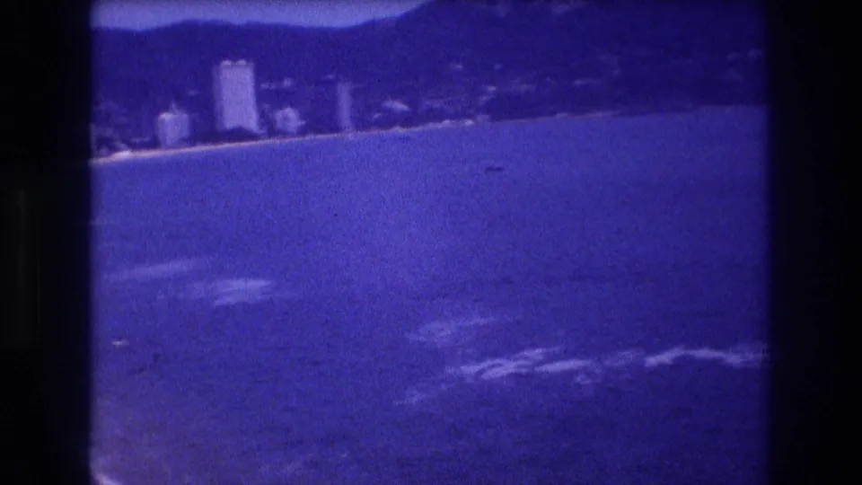 Final 8mm pan frame from Acapulco, Mexico, circa 1982: distant white high-rise hotels along a sandy beachfront with hazy foothills beyond.