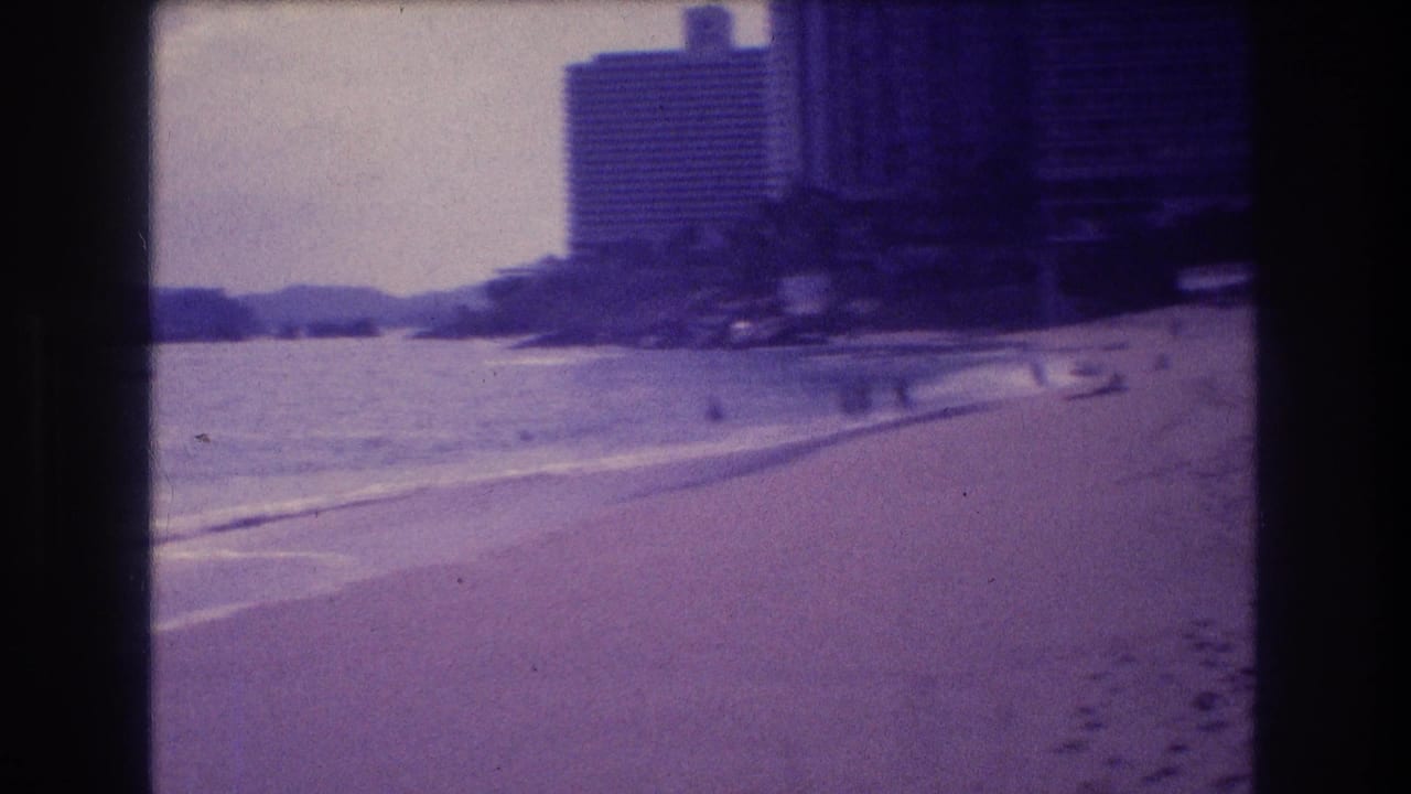 8mm frame from Acapulco c.1982 showing a sandy beach sweeping toward a high-rise hotel, with faint distant figures and a small object at the waterline.