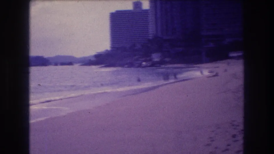 8mm frame from Acapulco c.1982 showing a sandy beach sweeping toward a high-rise hotel, with faint distant figures and a small object at the waterline.