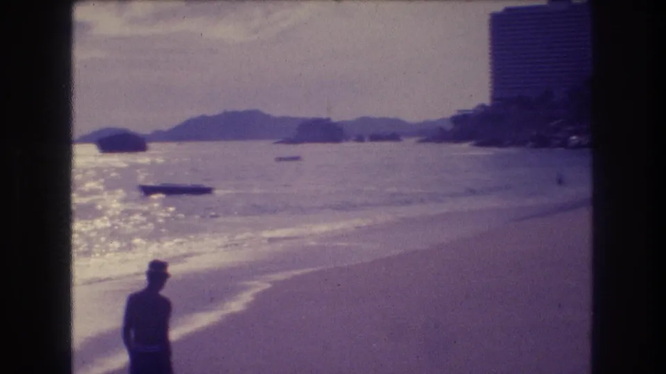 8mm home movie frame from Acapulco c.1982 — silhouetted figure at left, hotel tower behind, mountain ridgeline across the bay, small boats on shimmering water.