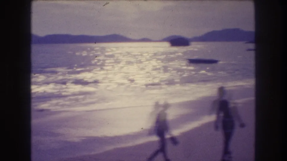 8mm home movie frame from Acapulco c.1982 — two motion-blurred figures walking near the waterline, mountain ridgeline across the bay, small boats visible on sun-lit water.