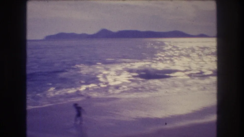 8mm home-movie frame showing a small figure at the water's edge on what appears to be an Acapulco beach, circa 1982, with sun glinting off the ocean and mountains in the distance.