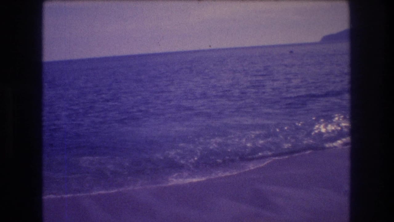 Sandy beach with a breaking wave at the shoreline and a mountainous headland visible at upper right, no figures clearly visible.