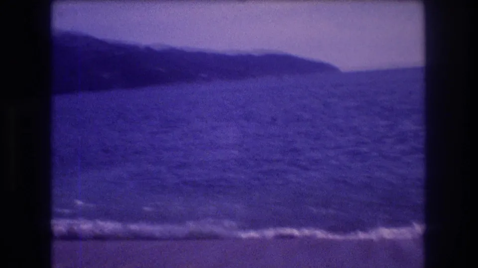 8mm home-movie wide shot of ocean waves and a dark hillside backdrop, possibly the Acapulco bay area, circa 1982, with a pronounced purple-blue color cast from aged film stock.