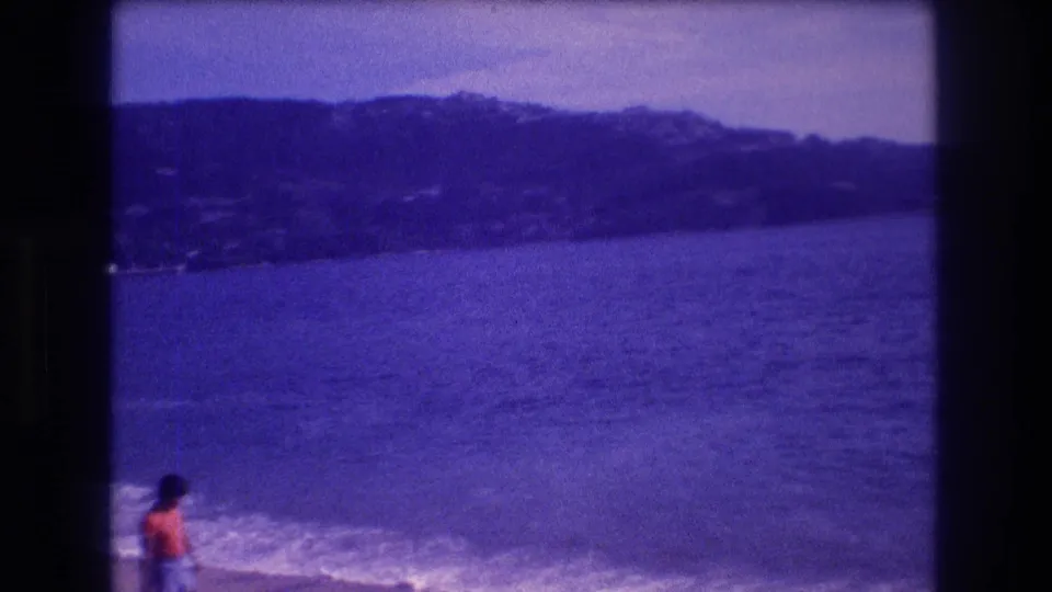 8mm home-movie frame of a small figure in bright clothing at the Acapulco shoreline, circa 1982, with a dark hillside showing possible resort development visible behind the bay.