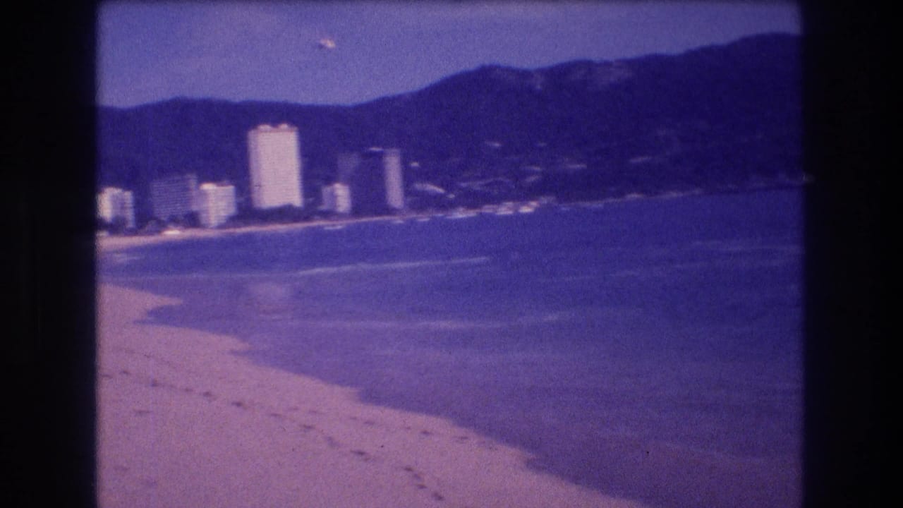 8mm home movie frame from Acapulco circa 1982 showing a sandy beach with footprints, whitecaps, a cluster of white high-rise hotels, and a mountain backdrop.