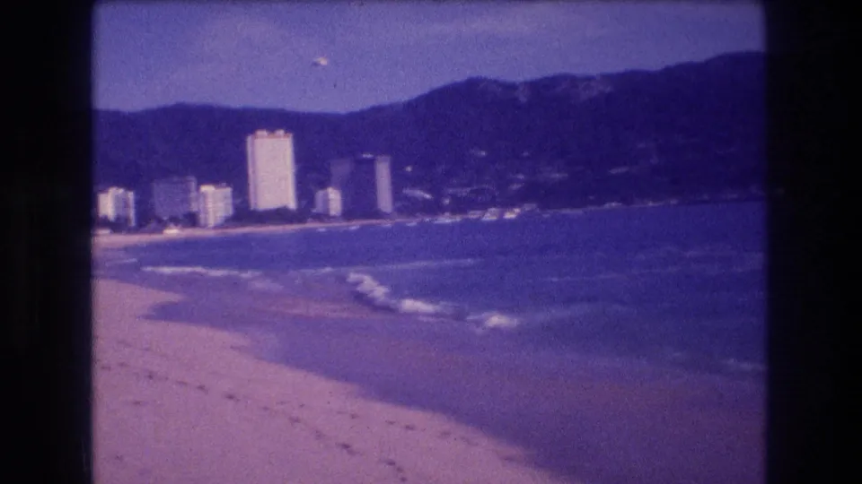 8mm home movie frame circa 1982 of the Acapulco coastline, with breaking surf, white hotel towers in mid-distance, and a ridgeline of dark mountains.
