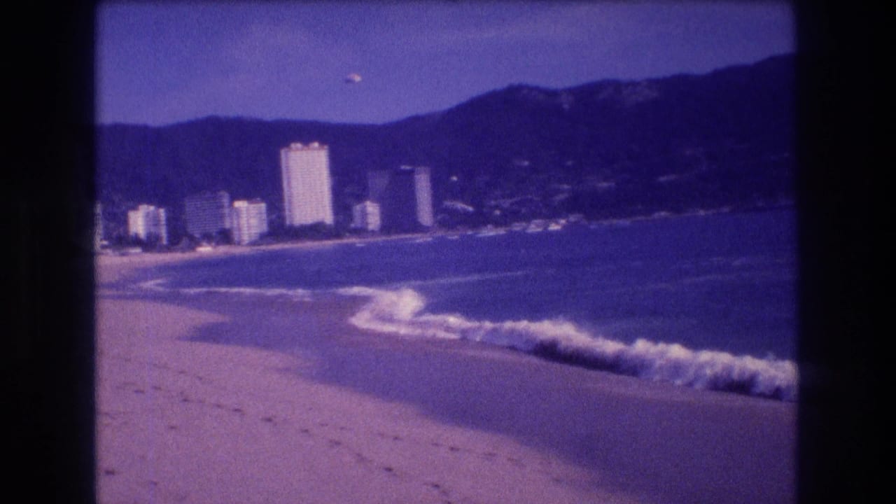 8mm home movie frame circa 1982 with more beach foreground in view, prominent whitecaps, distant Acapulco hotel towers, and dark mountains.