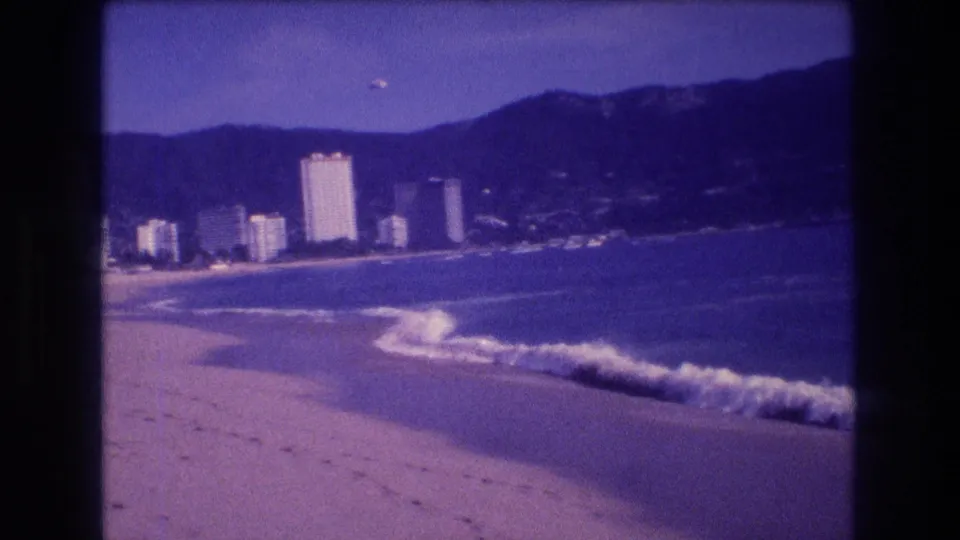 8mm home movie frame circa 1982 with more beach foreground in view, prominent whitecaps, distant Acapulco hotel towers, and dark mountains.
