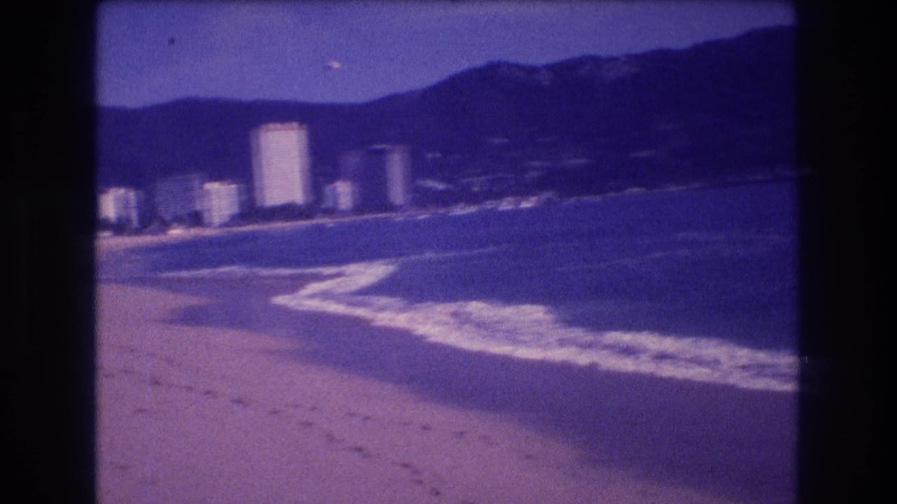 Late-clip 8mm frame from Acapulco circa 1982 showing beach sand, whitecaps curving along the shoreline, high-rise hotels in mid-distance, and a mountain ridge.