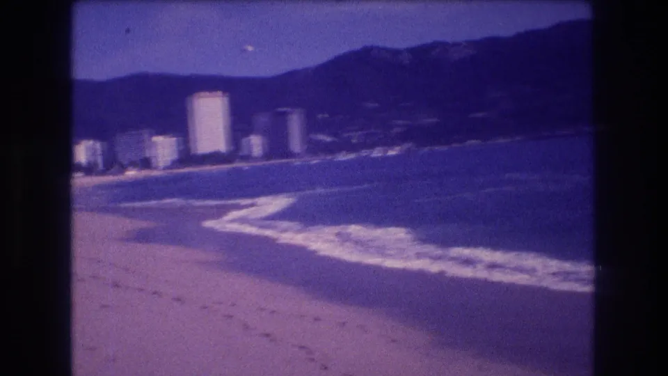 Late-clip 8mm frame from Acapulco circa 1982 showing beach sand, whitecaps curving along the shoreline, high-rise hotels in mid-distance, and a mountain ridge.
