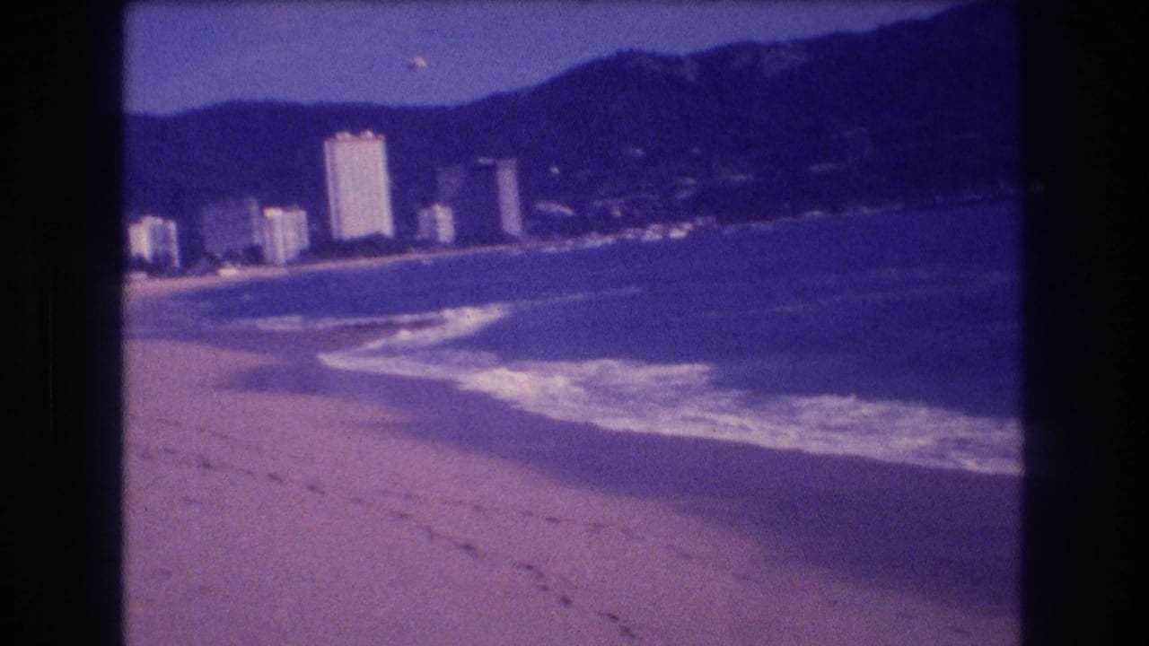 Sandy beach with footprints, breaking surf, and white hotel towers along an Acapulco bay backed by dark mountains.