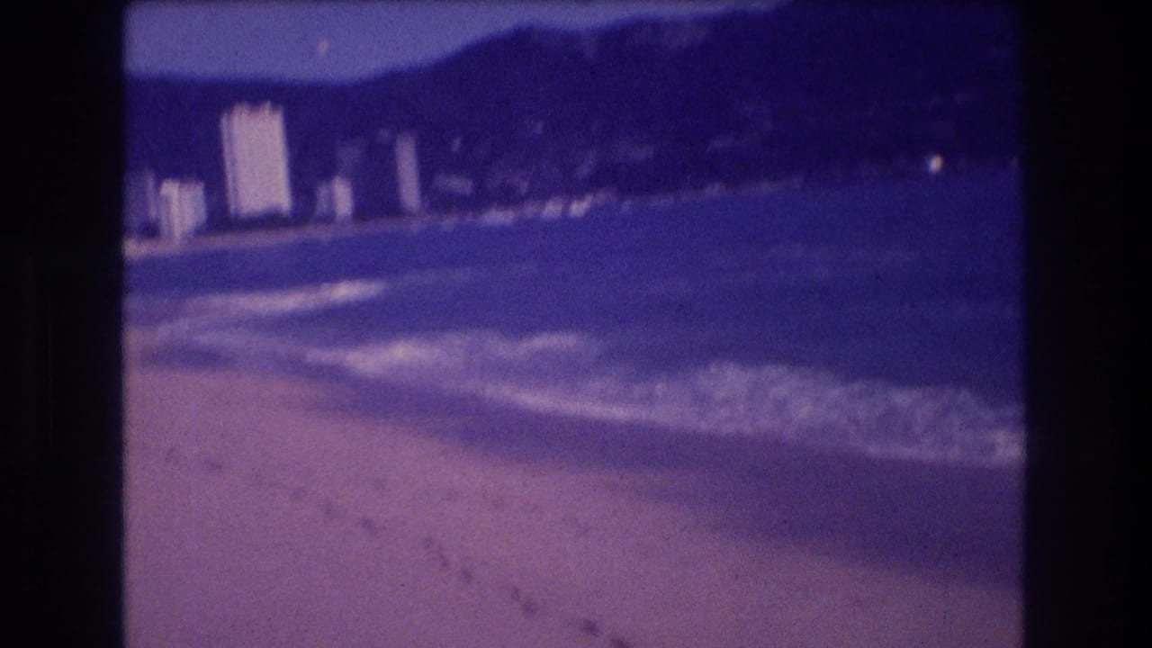 Acapulco beach and bay with white hotel towers at left, dark mountains behind, and wave foam along the curving shoreline.