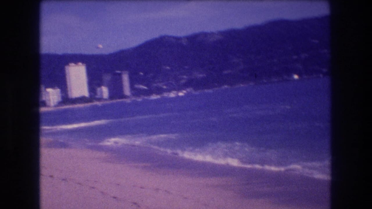 8mm home-movie frame showing a wider angle of Acapulco's bay, hotel cluster, and surrounding mountains under the characteristic aged-film blue-violet cast, circa 1982.